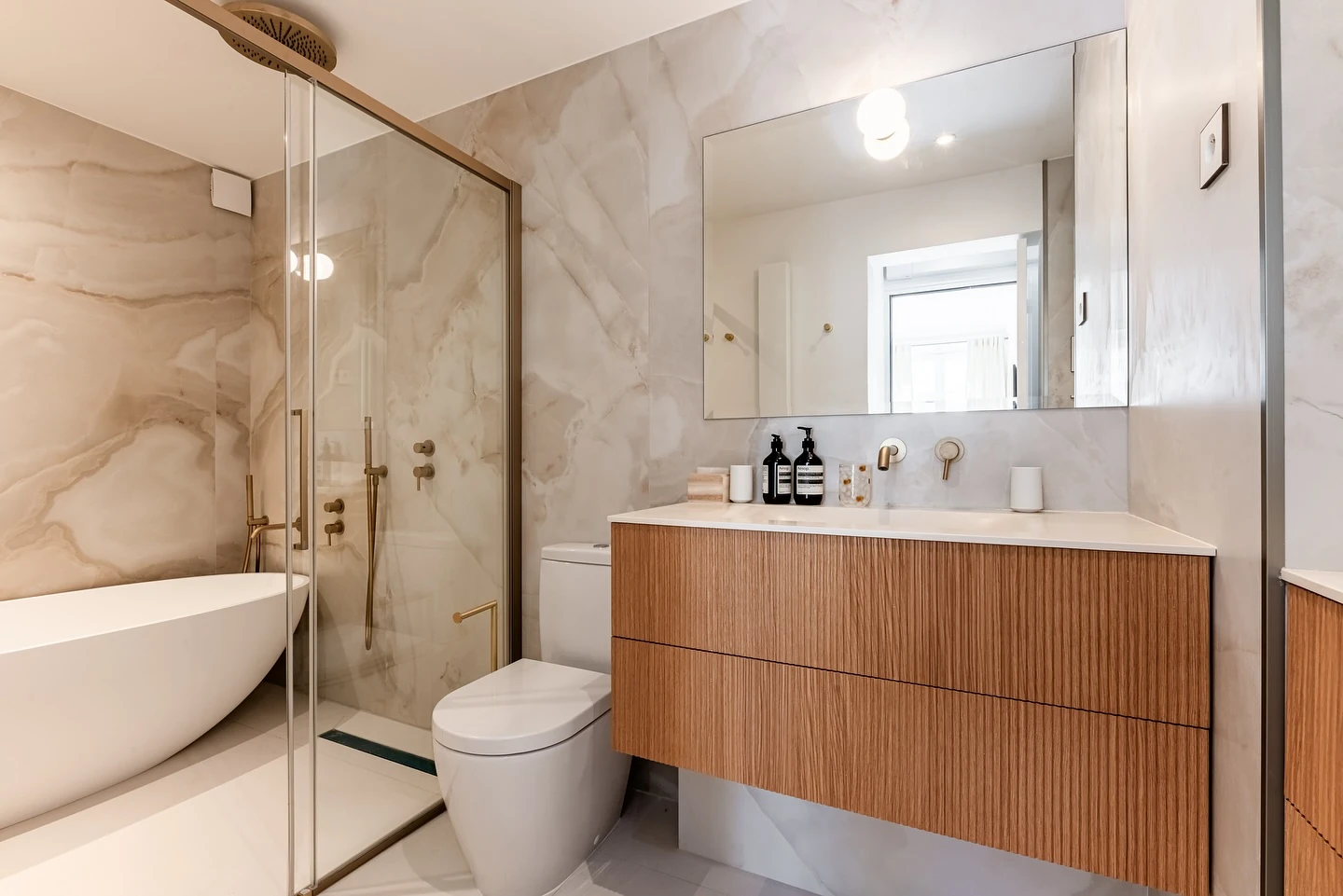 Salle de bain Odéon, revêtement clair veiné, meuble en bois et robinetterie en finition laiton.
